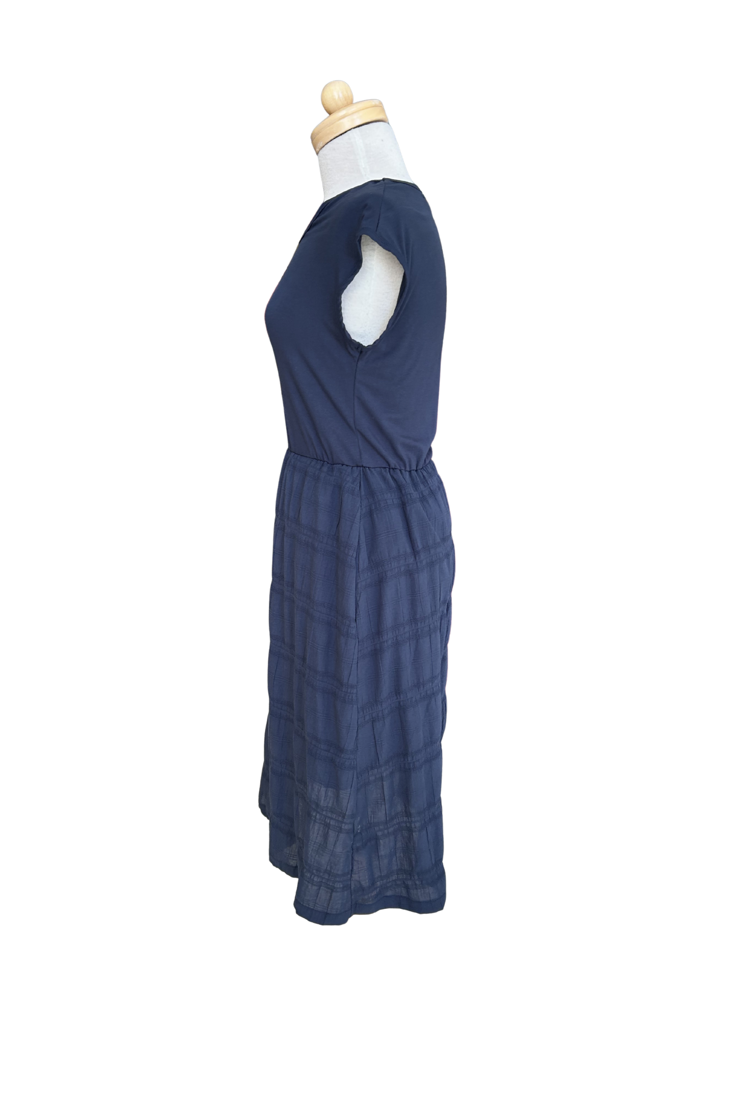 A side view of the Mona Dress by Tangente in Navy, a below the knee length dress featuring a bamboo rayon bodice and seersucker skirt, with short sleeves and a round neck, is shown on a mannequin against a white background. 
