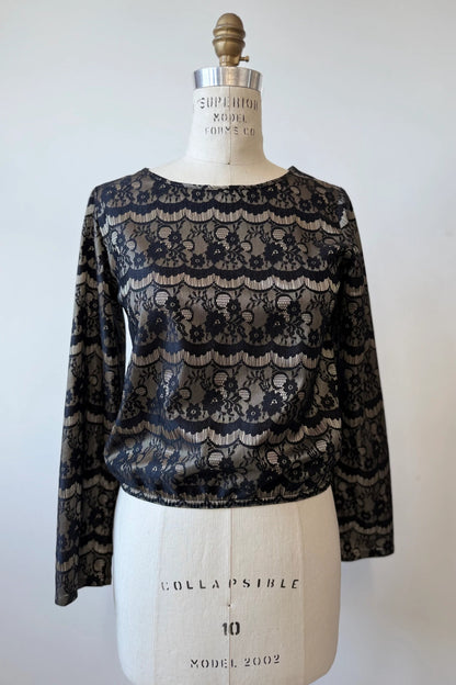 The Solstice Blouse by Kollontai in Gold is shown on a mannequin in front of a white background.  The fabric layers black lace over golden satin. It's slightly cropped with an elastic waist and long sleeves. 