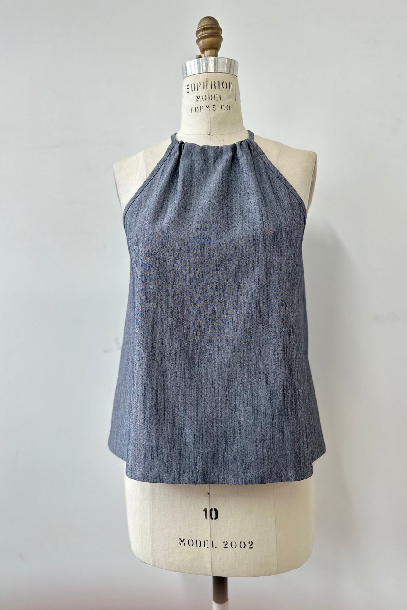 The Semira Camisole by Kollontai in denim, a lightweight top with an adjustable halter neck and a loose fit, is show on a mannequin in front of a white background. 