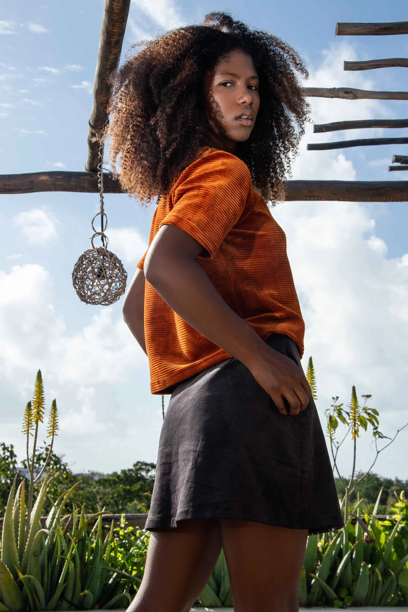 Side view of a woman wearing the Tofino Top by MAS in Amber, a slightly cropped tee in velvet rib. She is wearing it with a black skirt and is standing outside. 