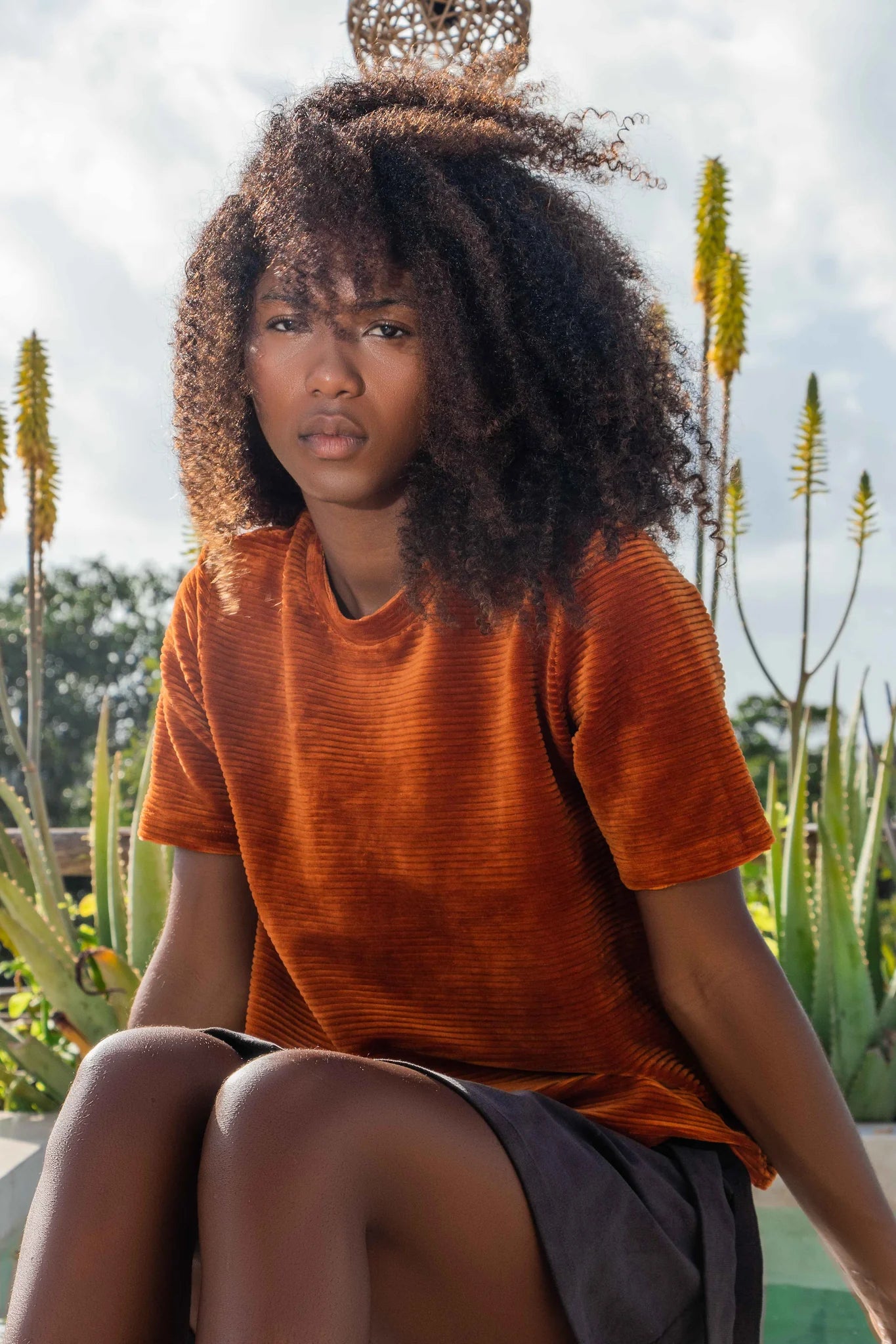 A woman wearing the Tofino Top by MAS in Amber, a slightly cropped tee in velvet rib. She is wearing it with a black skirt and is sitting in front of a garden.