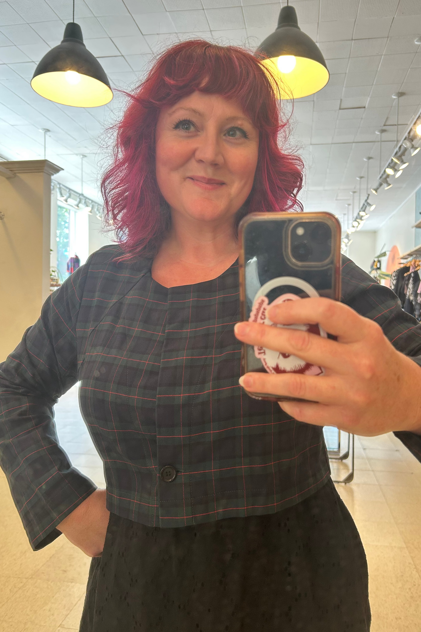 A woman with vibrant pink hair taking a  mirror selfie at Flock, wearing  the Libra Bolero by Allison Wonderland in Plaid, a green plaid cropped jacket with a single button closure. She is wearing it over a black dress.