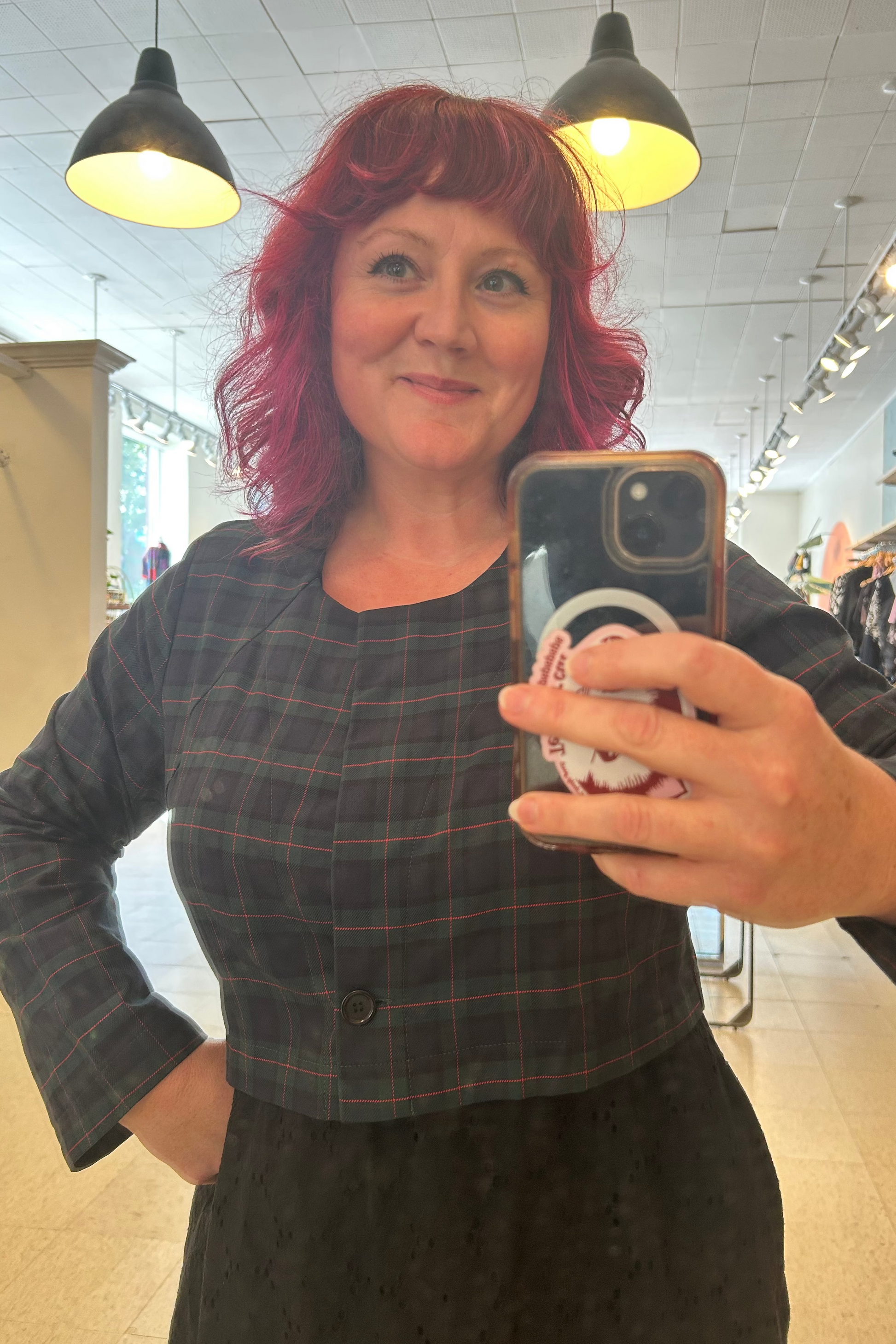 A woman with vibrant pink hair taking a  mirror selfie at Flock, wearing  the Libra Bolero by Allison Wonderland in Plaid, a green plaid cropped jacket with a single button closure. She is wearing it over a black dress.