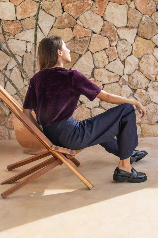 Side view of a woman wearing the Tofino Top by MAS in Amethyst, a slightly cropped tee in velvet rib. She is wearing it with a blue trousers and is sitting in a wooden chair. 