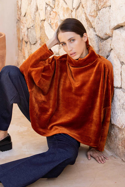 A woman wearing the Amsterdam Sweater by MAS in Amber, an oversized one-size top with a chimney collar, extended sleeves, and a cropped length, with blue trousers, sitting against a stone wall. 