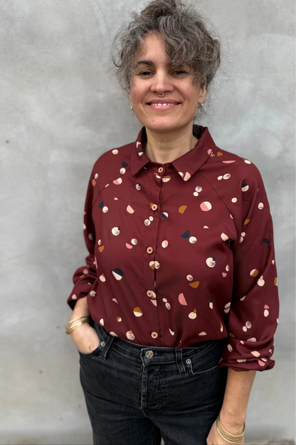 A woman wearing the Acer Blouse by Kazak in Burgundy Moons, a raglan sleeved blouse with a classic collar, button front, and elastic cuffs. She is wearing it with black jeans and standing in front of a grey background. 
