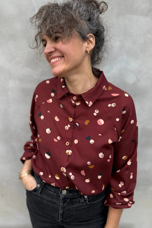 A woman wearing the Acer Blouse by Kazak in Burgundy Moons, a raglan sleeved blouse with a classic collar, button front, and elastic cuffs. She is wearing it with black jeans and standing in front of a grey background. 