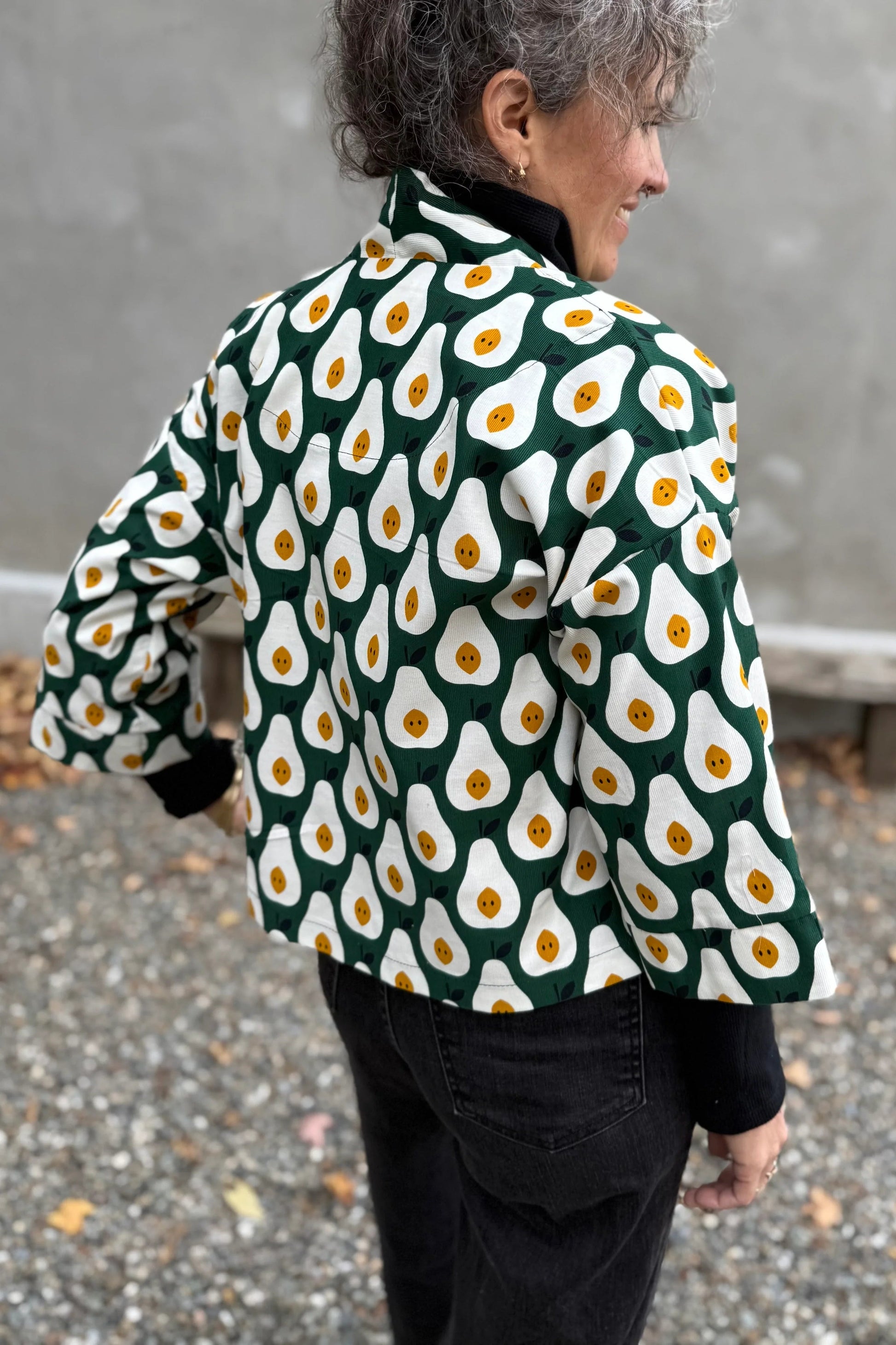Back view of a woman wearing the Magnolia Jacket by Kazak in green and white corduroy Pears print fabric, a short kimono style open jacket with 3/4 sleeves and large patch pockets. She is wearing it with a black top and black jeans and standing in front of a grey wall. 