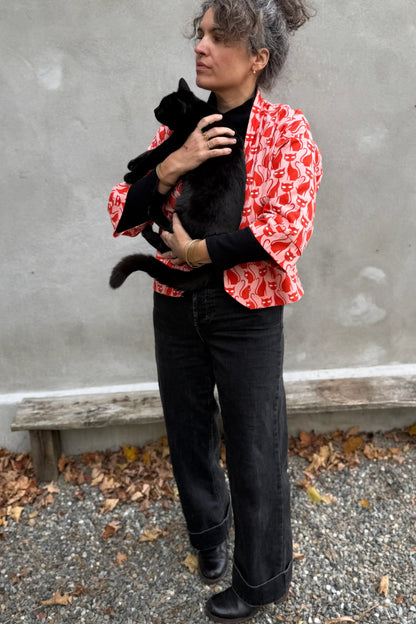 A woman wearing the Magnolia Jacket by Kazak in red corduroy Cats print fabric, a short kimono style open jacket with 3/4 sleeves and large patch pockets. She is wearing it with a black top and black jeans and holding a black cat while standing in front of a grey wall. 