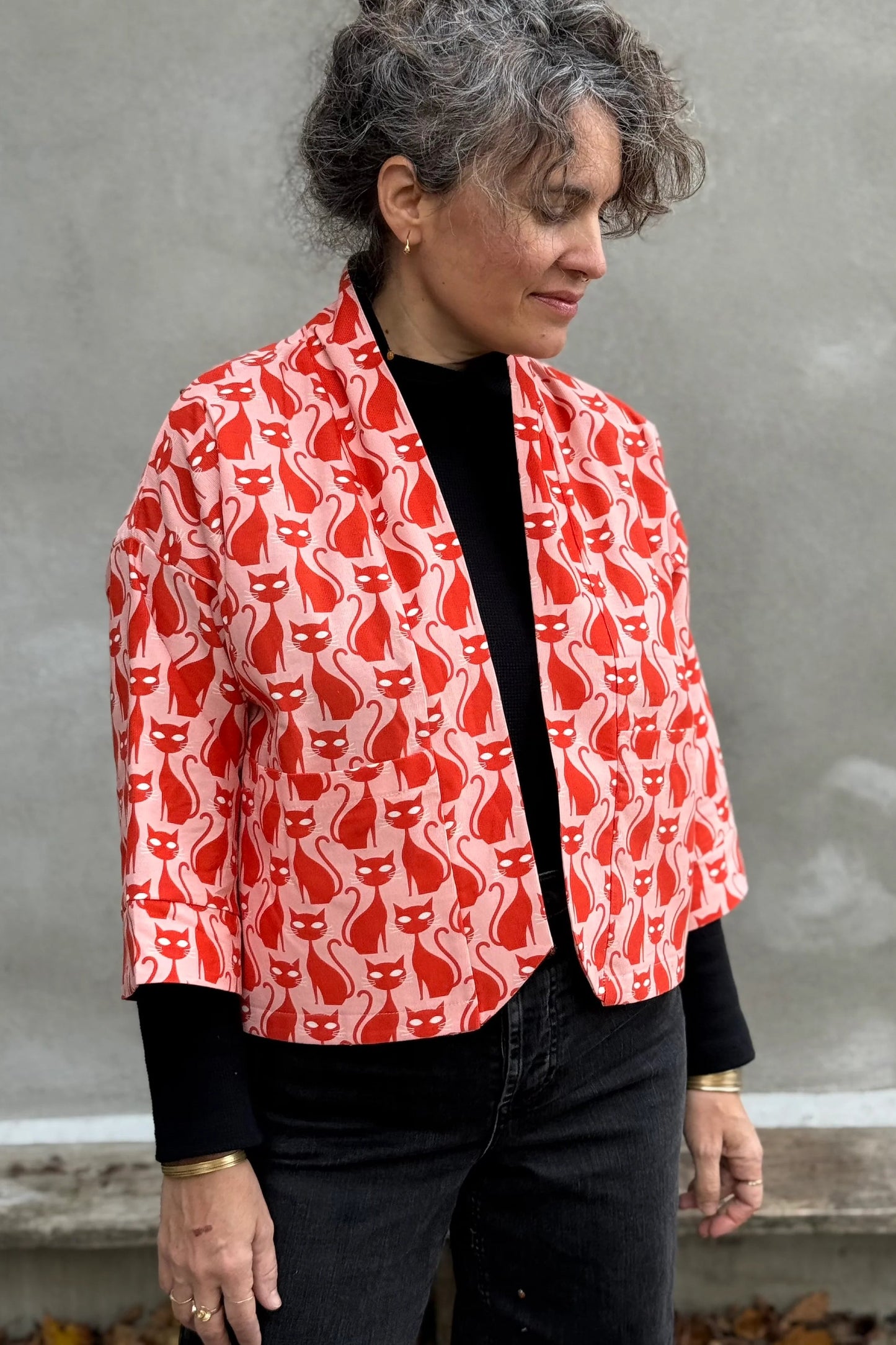 A woman wearing the Magnolia Jacket by Kazak in red corduroy Cats print fabric, a short kimono style open jacket with 3/4 sleeves and large patch pockets. She is wearing it with a black top and black jeans and standing in front of a grey wall. 