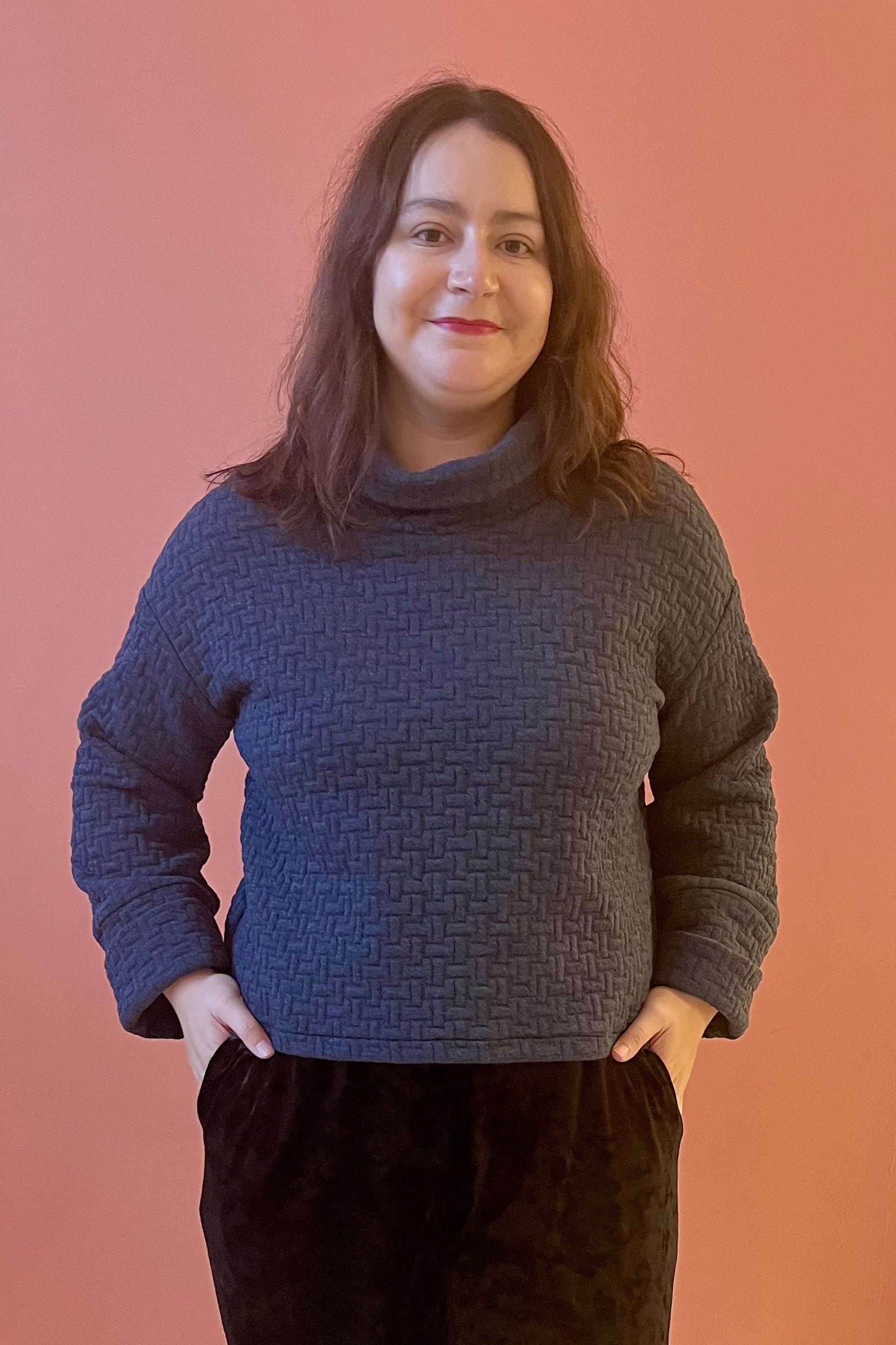 A woman wearing the Sable Top by Tangente in Dark Denim, a sweatshirt in quilted fabric with a cowl neck and cuffed sleeves and a slightly cropped and boxy fit. She is wearing it with black pants and standing in front of a coral background. 