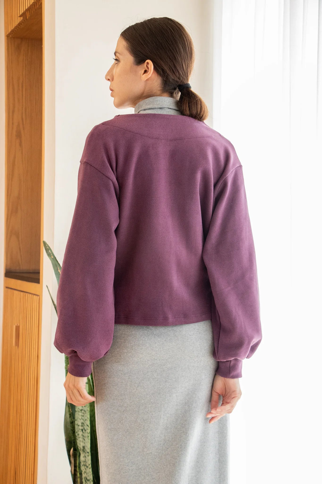 Back view of a woman wearing the Giza Sweater by MAS in Grape, with a two button closure and long sleeves with a puff detail at the cuff, over a grey dress. She is standing in front of a white curtain. 