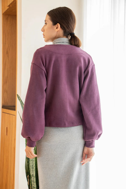 Back view of a woman wearing the Giza Sweater by MAS in Grape, with a two button closure and long sleeves with a puff detail at the cuff, over a grey dress. She is standing in front of a white curtain. 