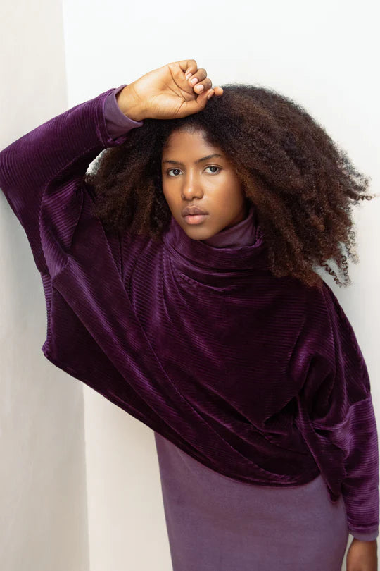 A woman wearing the Amsterdam Sweater by MAS in Amethyst, an oversized one-size top with a chimney collar, extended sleeves, and a cropped length, with a purple shirt underneath, leaning against a white wall. 