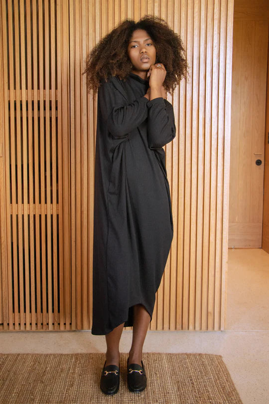 A woman wearing the Frankfurt Dress by MAS in Pepper, standing in front of a wooden wall
