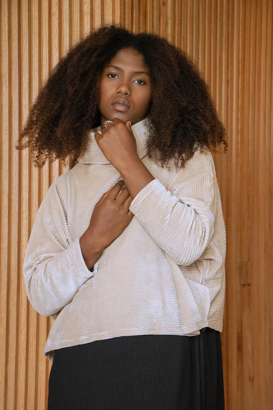 A woman wearing the Amsterdam Sweater by MAS in Latte, an oversized one-size top with a chimney collar, extended sleeves, and a cropped length, with blue trousers, leaning against a wooden wall. 