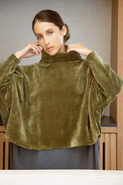 A woman wearing the Amsterdam Sweater by MAS in Moss, an oversized one-size top with a chimney collar, extended sleeves, and a cropped length, with a grey skirt, standing in front of a wooden table. 