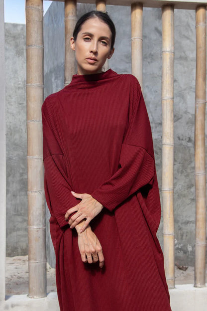 A woman wearing the Frankfurt Dress by MAS in Ruby, standing outside front of a bamboo fence 