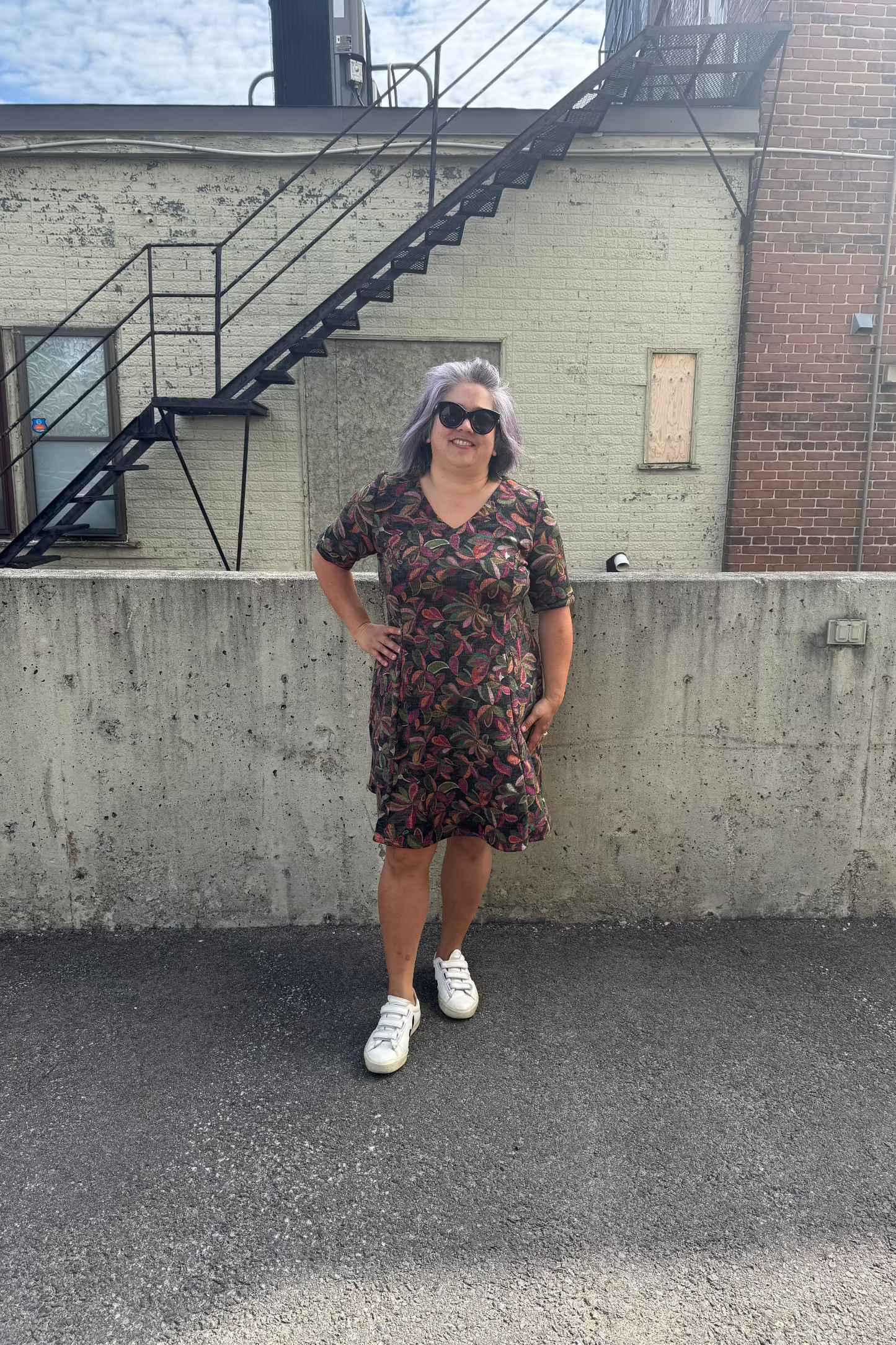 A woman wearing the Botanical Dress by Karkass in Floral, a knit dress with a V-neck, elbow length sleeves, fit and flare shape, and above the knee hem.  She is standing in front of a building with a fire escape.  