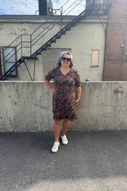 A woman wearing the Botanical Dress by Karkass in Floral, a knit dress with a V-neck, elbow length sleeves, fit and flare shape, and above the knee hem.  She is standing in front of a building with a fire escape.  