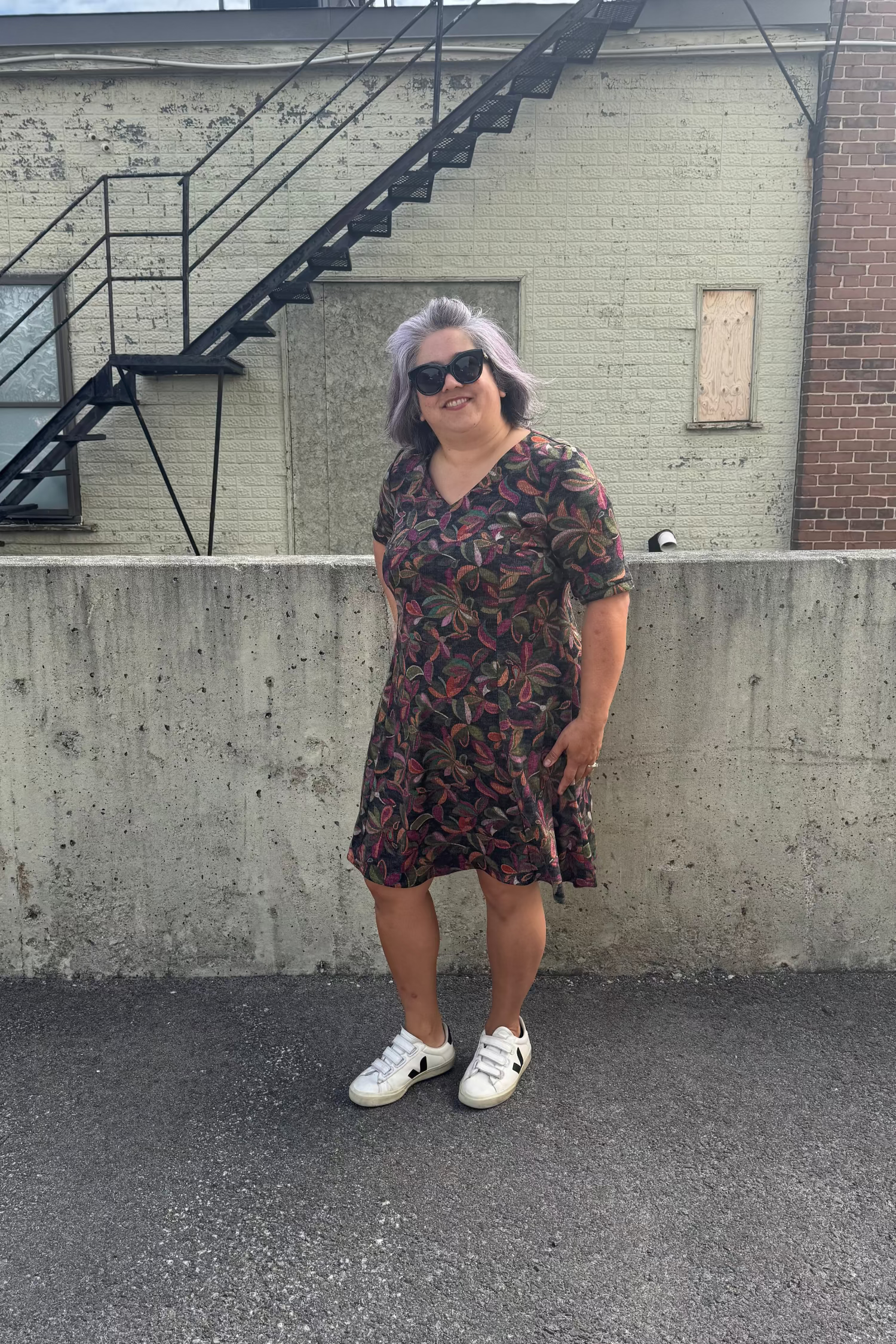 A woman wearing the Botanical Dress by Karkass in Floral, a knit dress with a V-neck, elbow length sleeves, fit and flare shape, and above the knee hem.  She is standing in front of a building with a fire escape.  