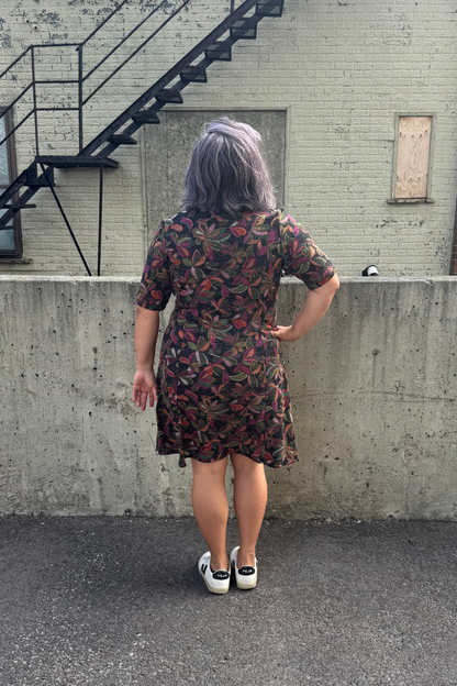 Back view of a woman wearing the Botanical Dress by Karkass in Floral, a knit dress with a V-neck, elbow length sleeves, fit and flare shape, and above the knee hem.  She is standing in front of a building with a fire escape.  