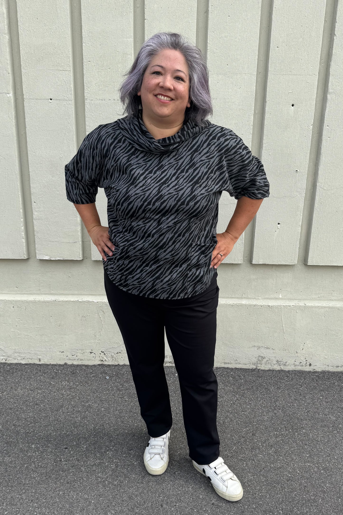 A woman wearing the Francis Tunic by Pure in Black/Charcoal animal print, featuring a turtleneck, 3/4 sleeves and two patch pockets. She is wearing it with Black pants and standing in front of a grey wall. 