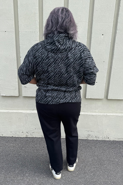 Back view of a woman wearing the Francis Tunic by Pure in Black/Charcoal animal print, featuring a turtleneck, 3/4 sleeves and two patch pockets. She is wearing it with Black pants and standing in front of a grey wall. 