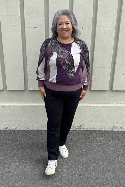 A woman wearing the Erin Sweater by Solomia in Cream/Black/Purple, a colour blocked top with a round neck and long sleeves, and a mix of patterns an solid colours including purple leopard print. She is wearing it with black pants and standing outdoors in front of a grey wall. 