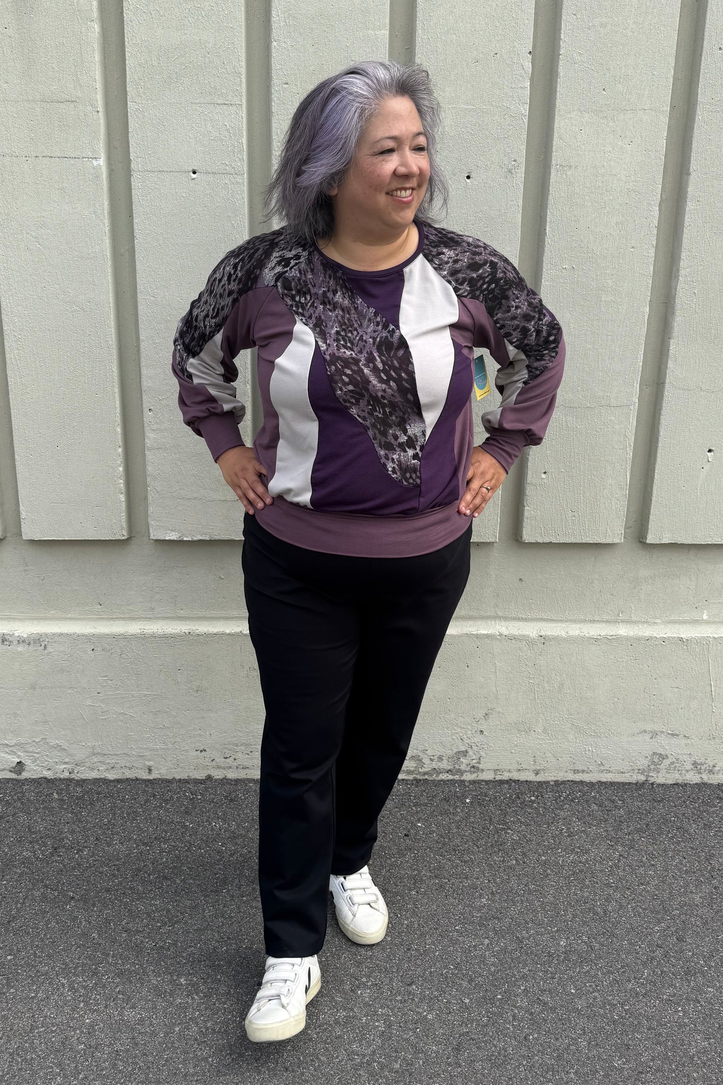 A woman wearing the Erin Sweater by Solomia in Cream/Black/Purple, a colour blocked top with a round neck and long sleeves, and a mix of patterns an solid colours including purple leopard print. She is wearing it with black pants and standing outdoors in front of a grey wall. 