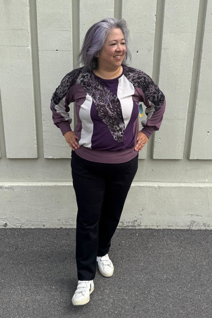 A woman wearing the Erin Sweater by Solomia in Cream/Black/Purple, a colour blocked top with a round neck and long sleeves, and a mix of patterns an solid colours including purple leopard print. She is wearing it with black pants and standing outdoors in front of a grey wall. 