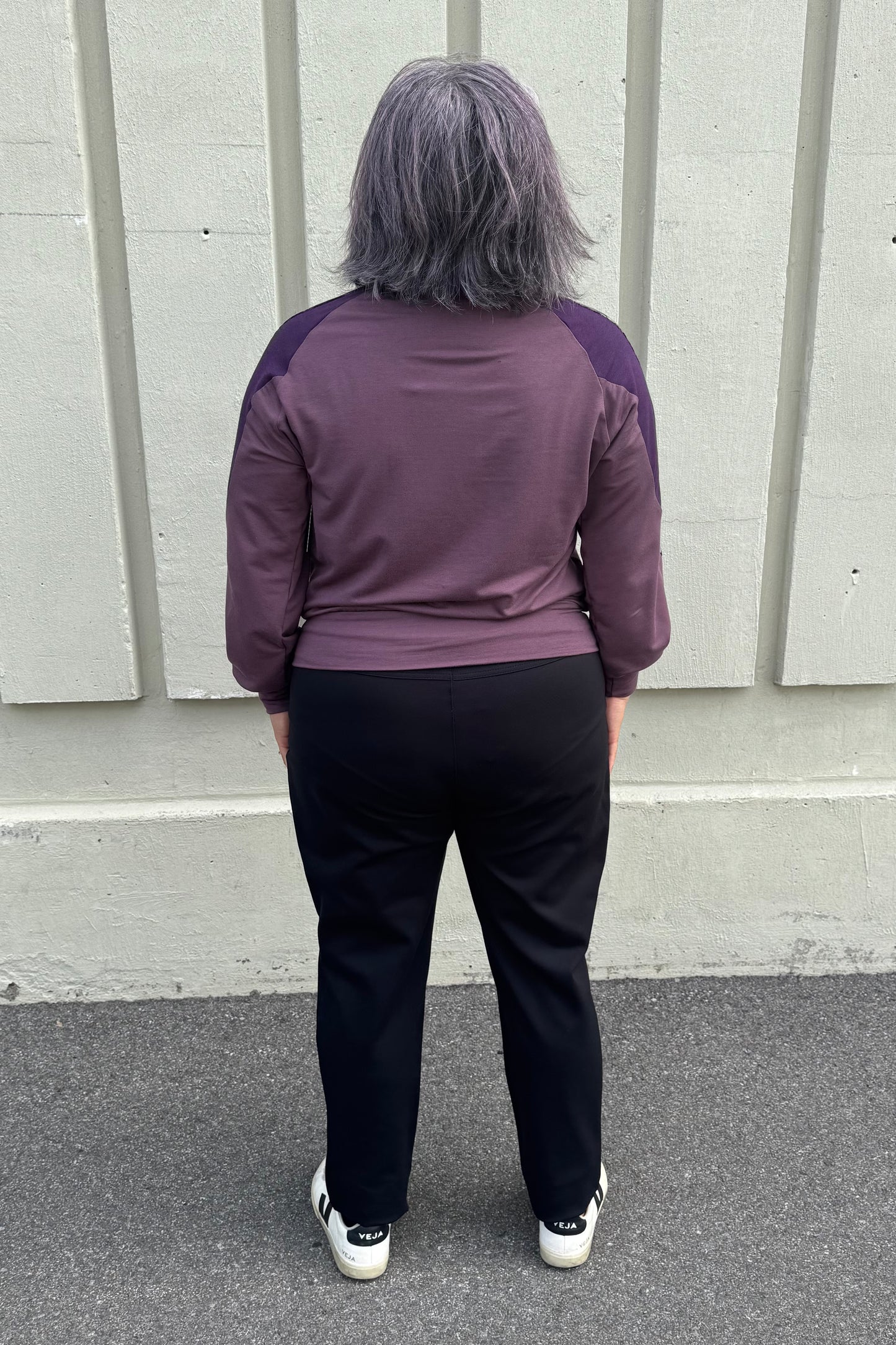 Back view of a woman wearing the Erin Sweater by Solomia in Cream/Black/Purple, a colour blocked top with a round neck and long sleeves, and a mix of patterns an solid colours including purple leopard print. She is wearing it with black pants and standing outdoors in front of a grey wall. 