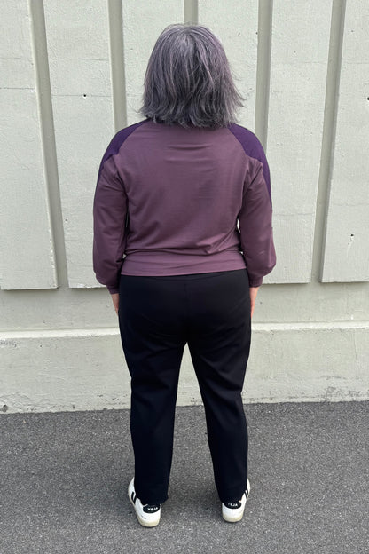 Back view of a woman wearing the Erin Sweater by Solomia in Cream/Black/Purple, a colour blocked top with a round neck and long sleeves, and a mix of patterns an solid colours including purple leopard print. She is wearing it with black pants and standing outdoors in front of a grey wall. 