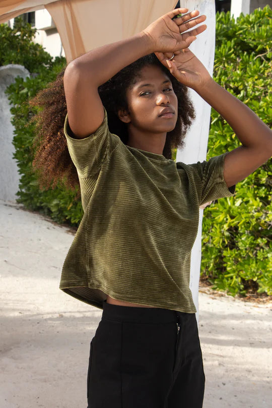 A woman wearing the MAS Tofino Top in Moss, a velvet rib t-shirt with a round neck, short sleeves, and a slightly cropped length. She is wearing it with black pants and standing in front of a bush. 