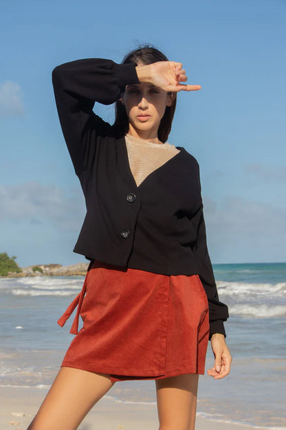 A woman wearing the Giza Sweater by MAS in Pepper, with a two button closure and long sleeves with a puff detail at the cuff, with a red skirt. She is standing in front of the ocean. 