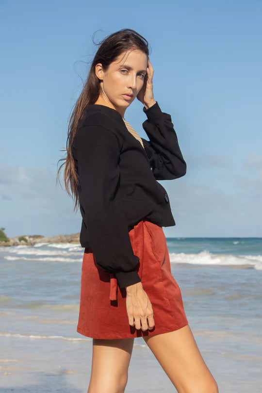 Side view of  woman wearing the Giza Sweater by MAS in Pepper, with a two button closure and long sleeves with a puff detail at the cuff, with a red skirt. She is standing in front of the ocean. 