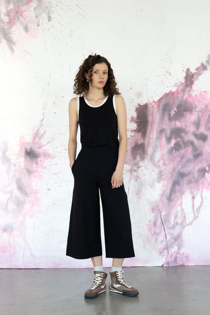 A woman wearing the Trille Trousers by Eve lavoie in Burgundy, wide legged 3/4 length pants with a wide pull-on waistband. She is wearing them with a black tank and standing in front of a speckled purple background. 