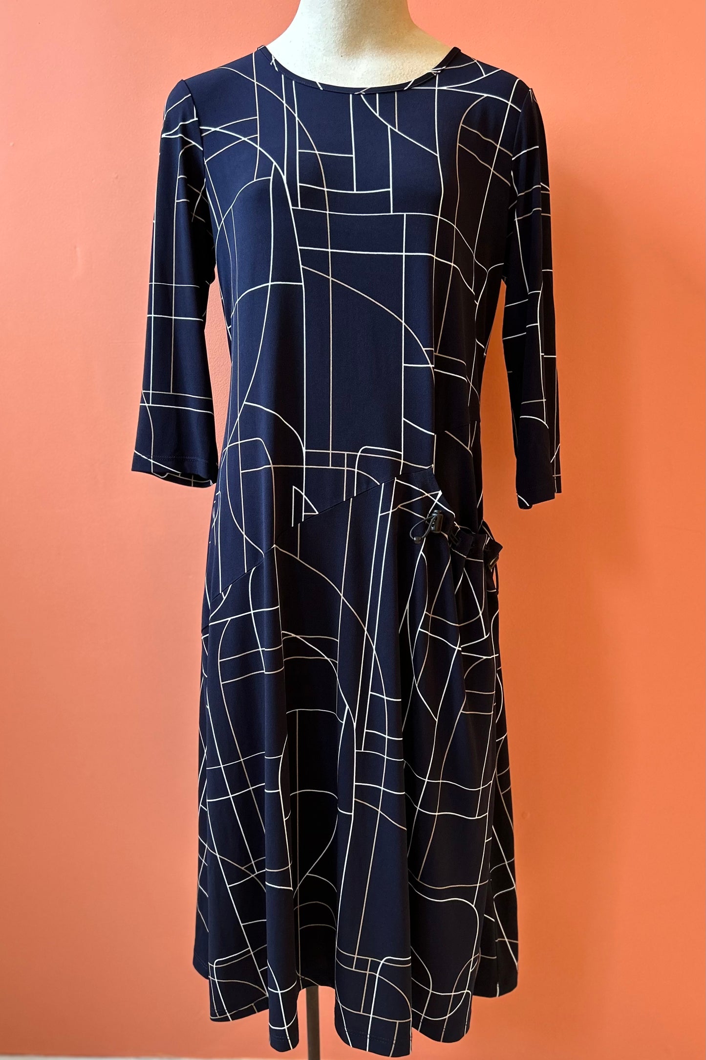 The Stormy Dress by Compli K in a navy geometric print is shown on a mannequin in front of a coral background