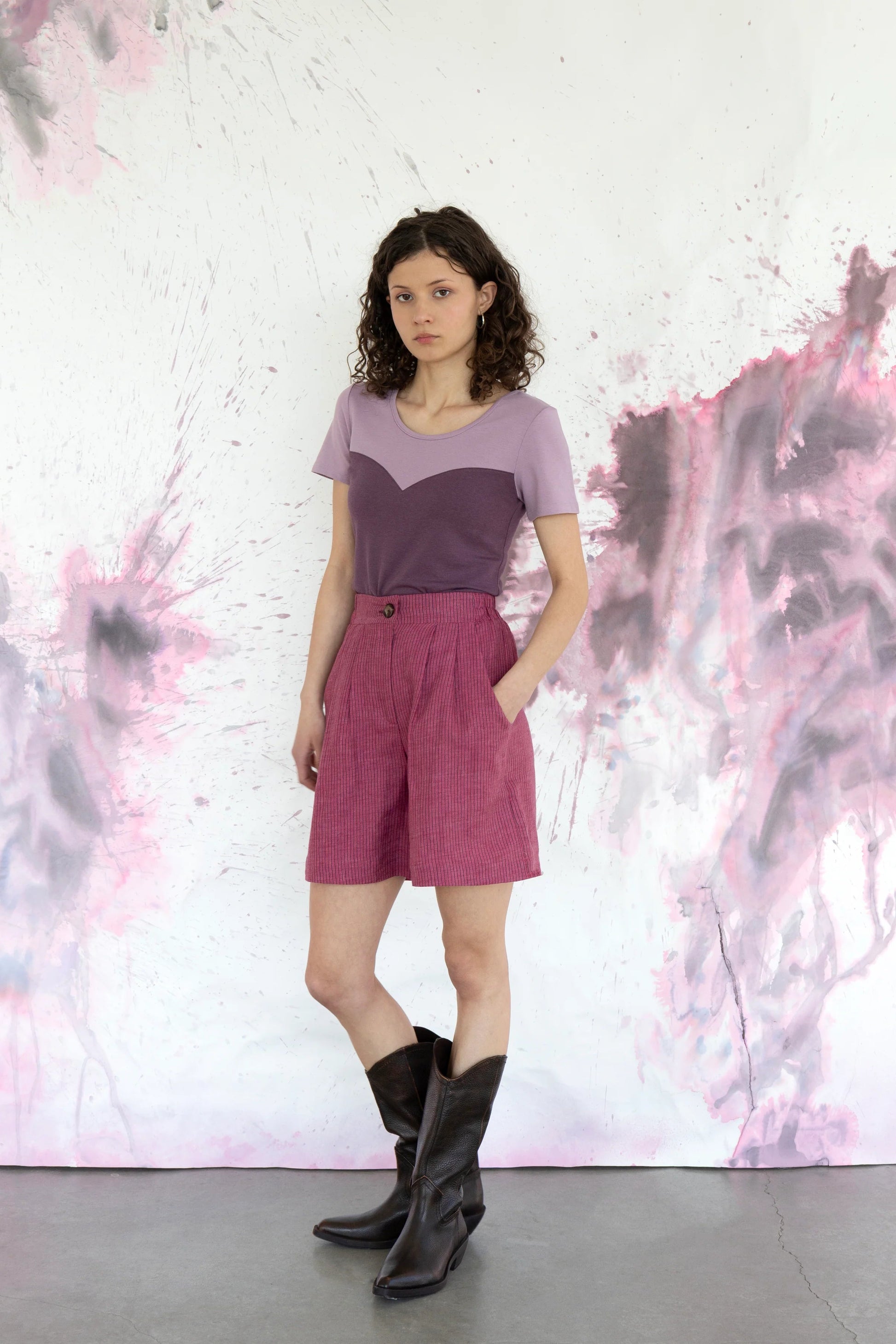 A woman wearing the Aster Shorts by Eve Lavoie in Raspberry Stripe with an 8-inch inseam, double pleats, a zip fly, an elasticated waistband and side pockets. She is wearing them with the Gentiane Top in Grape/Lilac and standing in front of a purple and white speckled background.