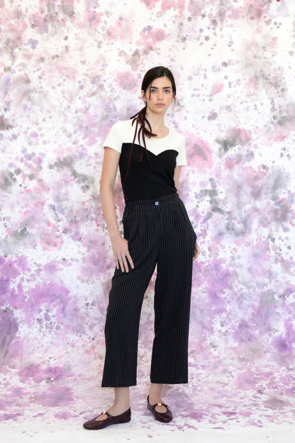 A woman wearing the Tanaisie Woven Pants from Eve Lavoie in Black Stripe, slightly cropped linen/cotton pants with wide legs, an elastic waist, classic fly closure, and slanted side pockets. She is wearing them with a black and white top and standing in front of a purple and white speckle background. 