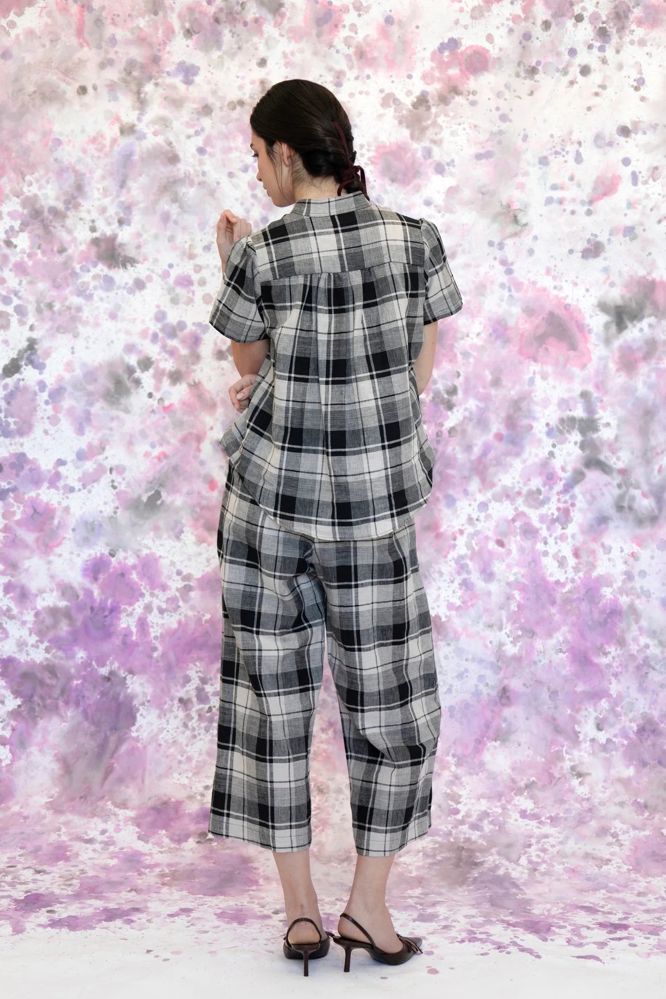 A woman wearing the Tanaisie Woven Pants from Eve Lavoie in Black and Cream Plalid, slightly cropped linen/cotton pants with wide legs, an elastic waist, classic fly closure, and slanted side pockets. She is wearing them with a black and white top and standing in front of a purple and white speckle background. 