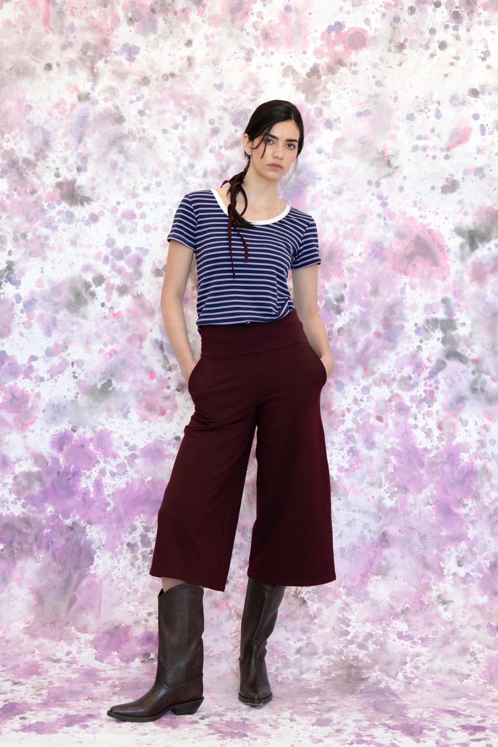 A woman wearing the Trille Trousers by Eve lavoie in Burgundy, wide legged 3/4 length pants with a wide pull-on waistband. She is wearing them with a blue striped t-shirt and standing in front of a speckled purple background. 