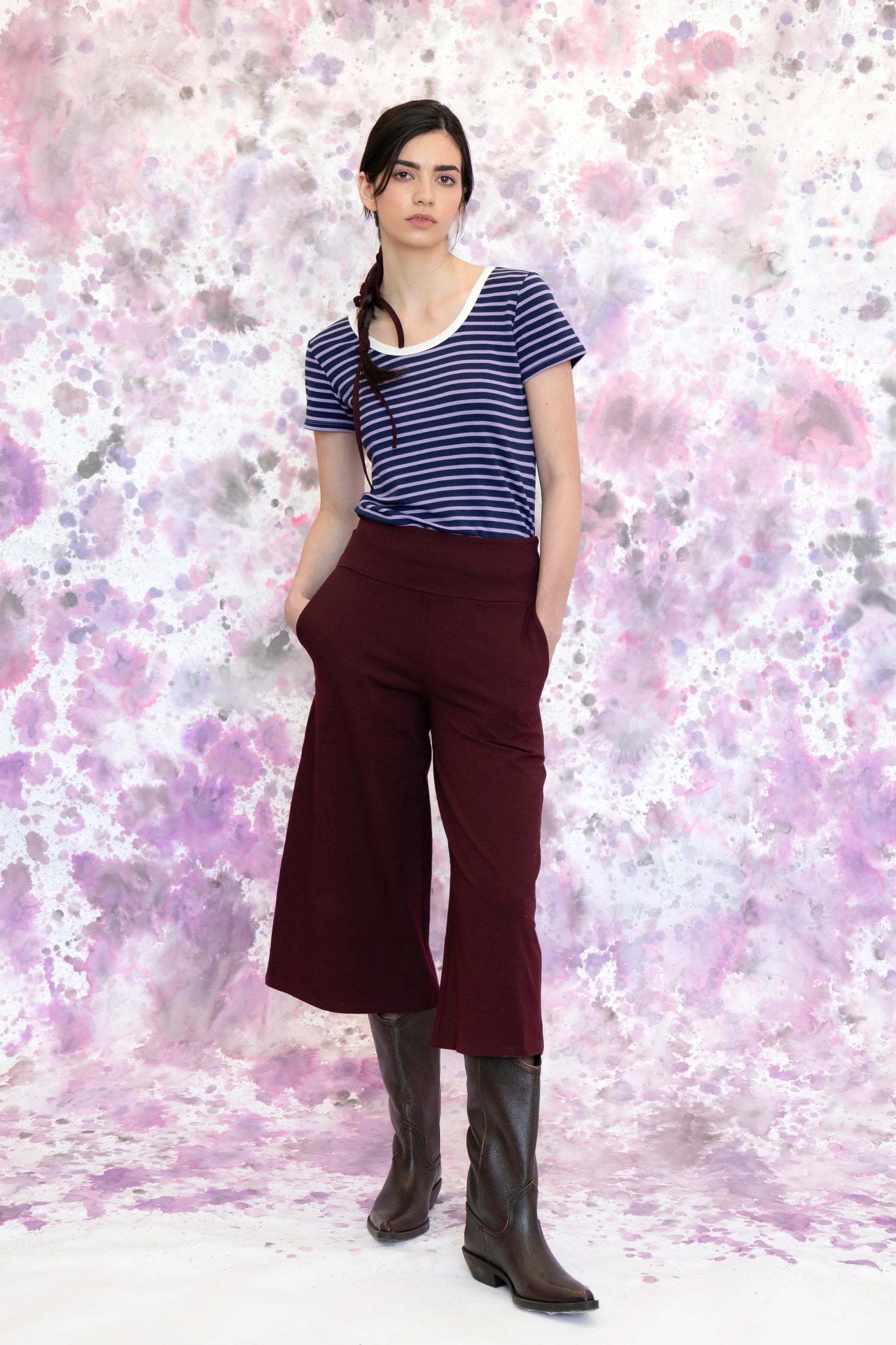 A woman wearing the Monarde Top by Eve Lavoie in Lavender Stripe with white piping. It is reversible back to front, with U-neckline on one side and a boat neckline on the other. She is wearing it with the Trille Trousers in  Burgundy pants and standing against an abstract background