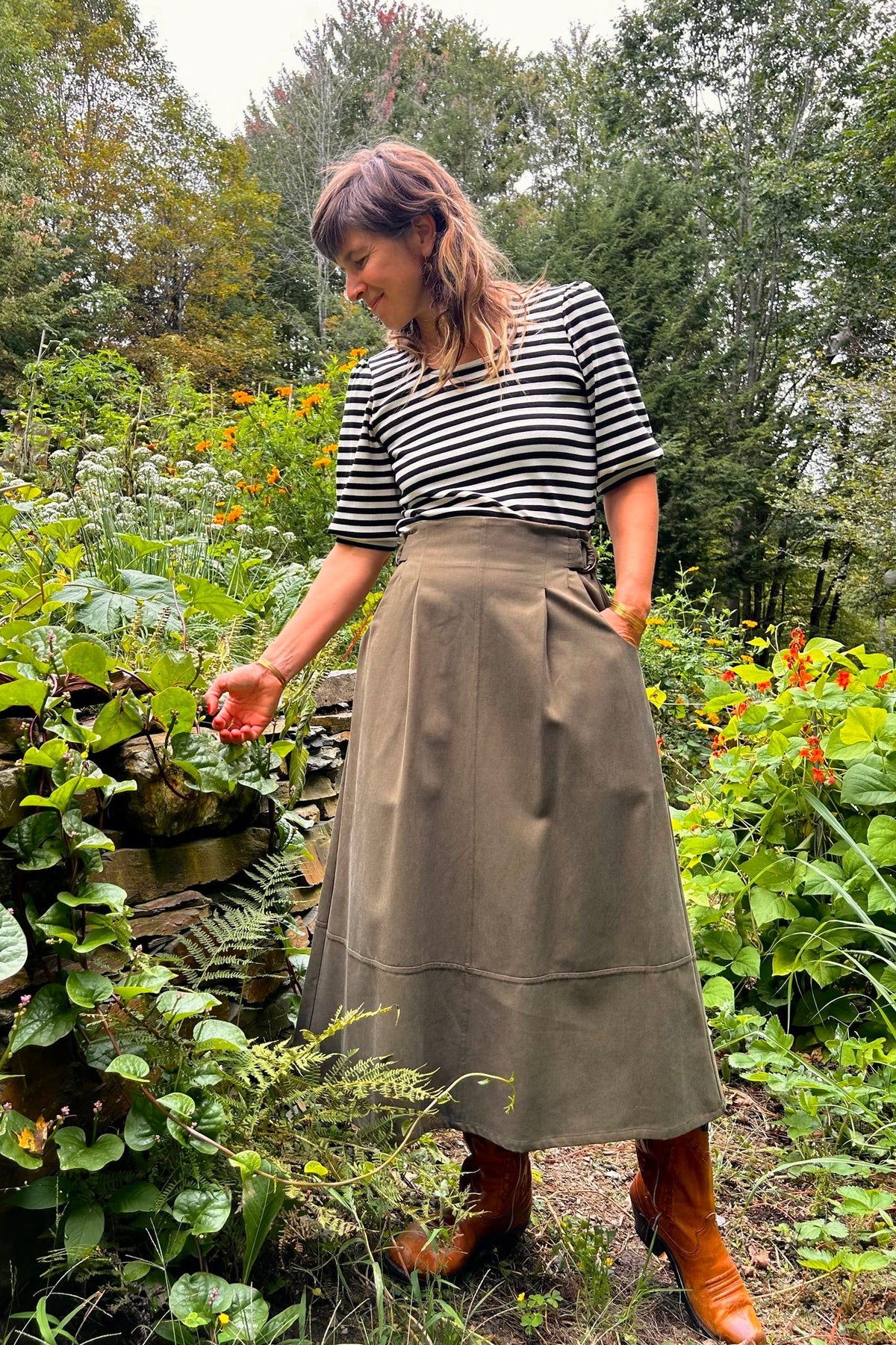 A woman standing in a lush garden wearing a black and white striped short-sleeve top, Kazak's Asarum skirt in Olive – a high-waisted midi skirt – and brown leather boots.
