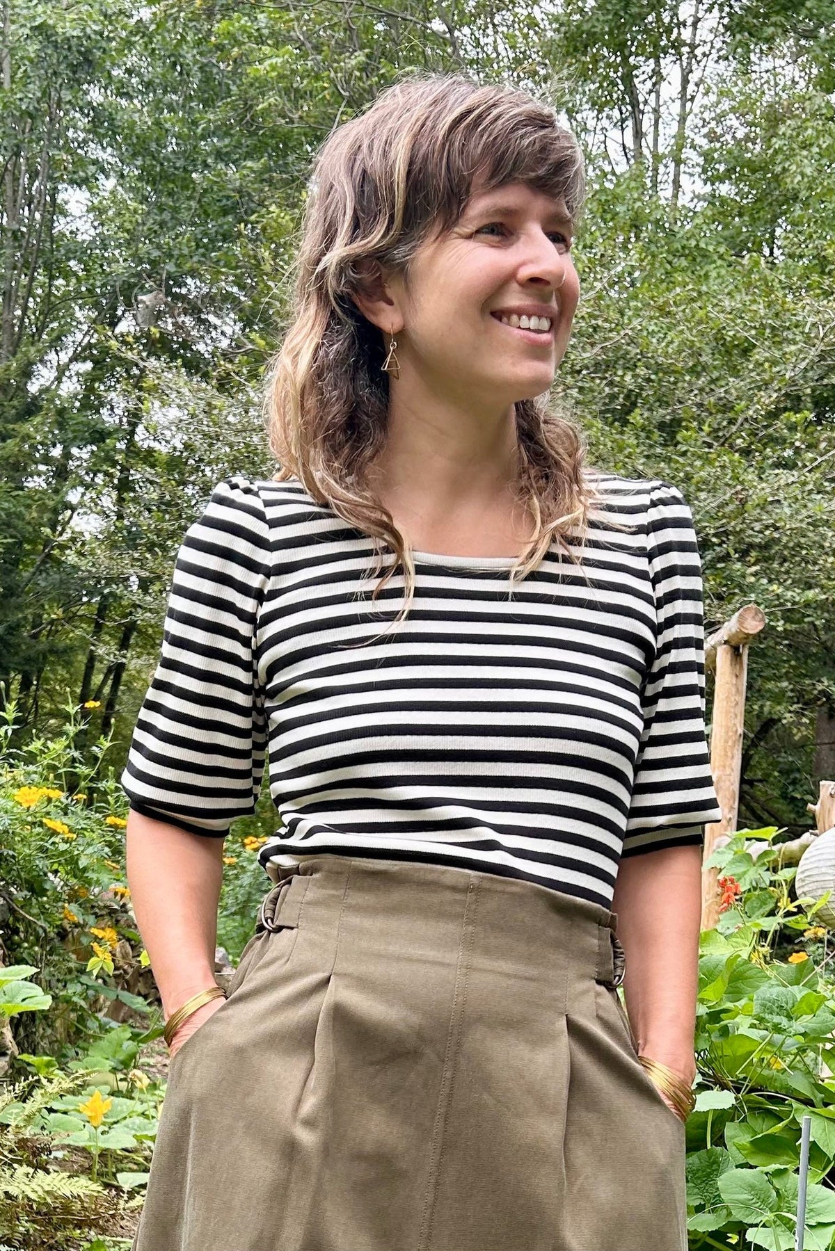 A woman standing in a lush garden wearing a black and white striped short-sleeve top, Kazak's Asarum skirt in Olive – a high-waisted midi skirt – and brown leather boots.