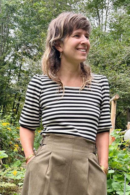 A woman standing in a lush garden wearing a black and white striped short-sleeve top, Kazak's Asarum skirt in Olive – a high-waisted midi skirt – and brown leather boots.