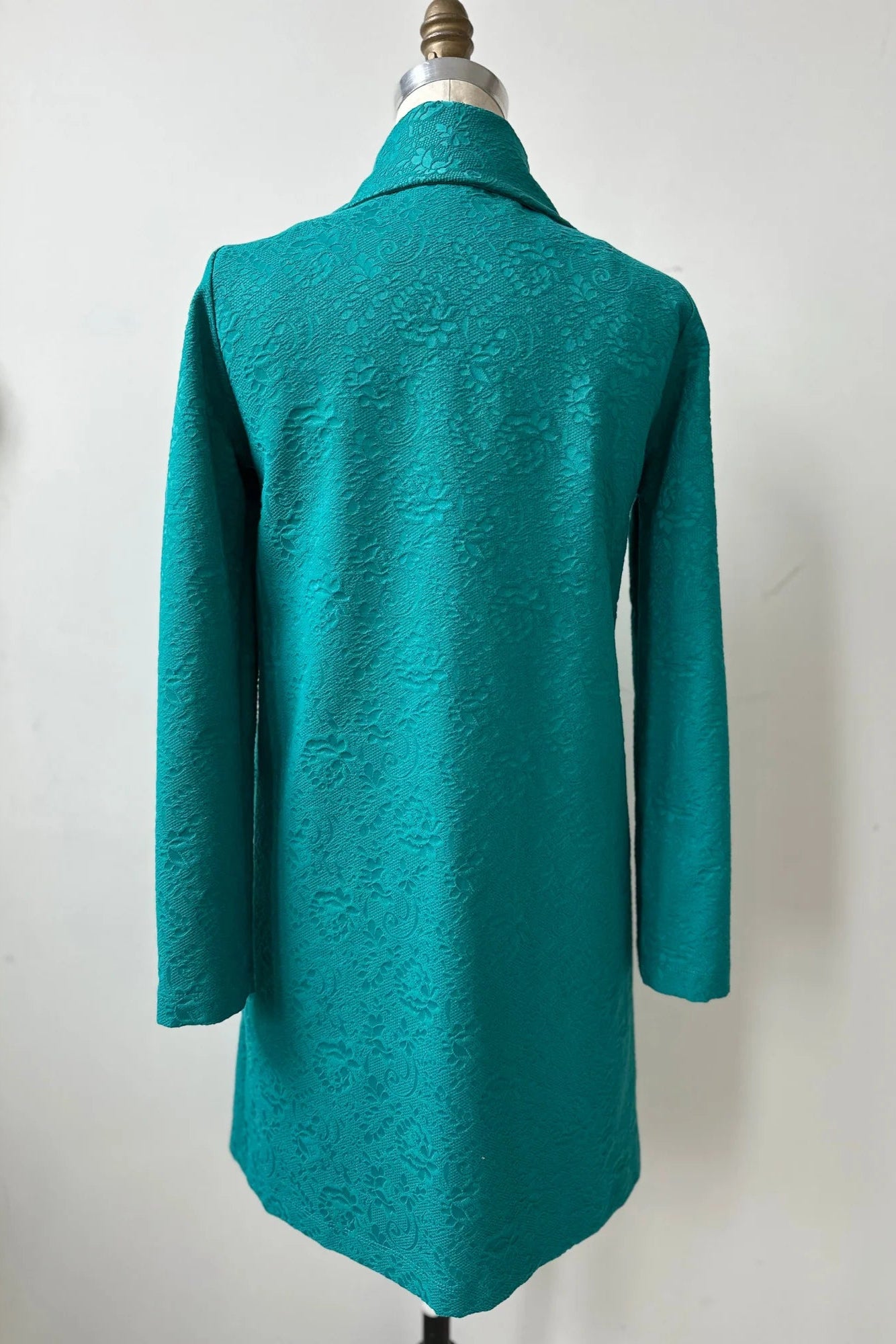 The back of the Emmanuelle Coat by Kollontai in Turquoise jacquard fabric is shown on a mannequin in front of a white background. It has a straight cut, classic collar, large patch pockets, and falls just above the knee. 