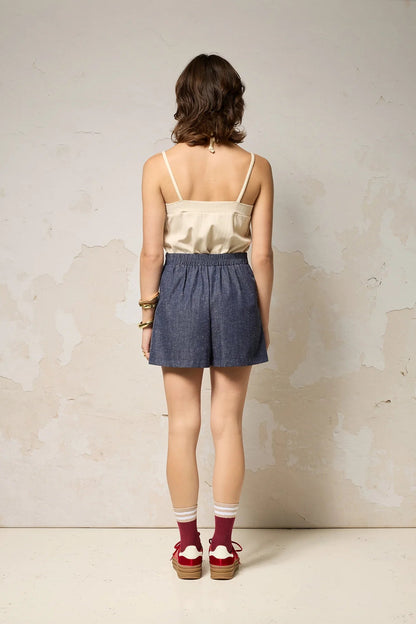 Back view of a woman wearing the Maya shorts from Cokluch in Indigo Speckle, lightweight denim shorts with a waist that's elasticized at the back, wide culotte style legs, and a side pockets. She is wearing them with a white tank and standing in front of a grey background. 