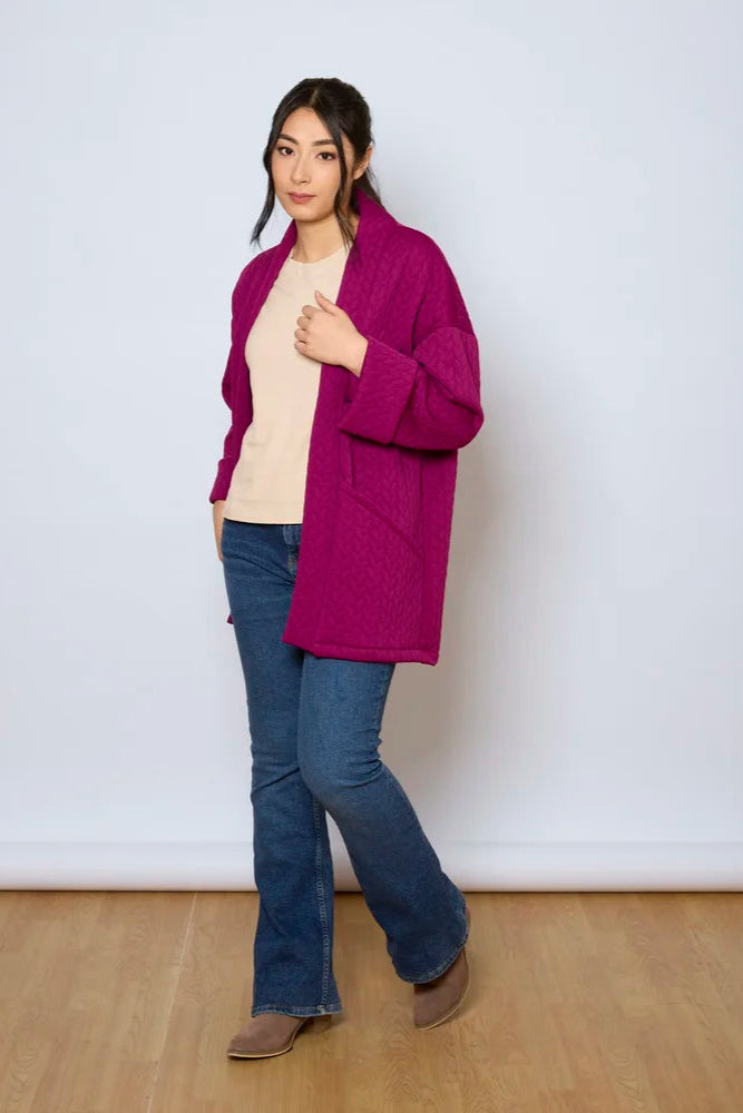 A woman wearing the Ina Coatigan by Tangente in Magenta, an open front cardigan in a cable-knit textured fabric, with dropped sleeves and patch pockets. She is wearing it with a white shirt and jeans and standing in front of a white background. 