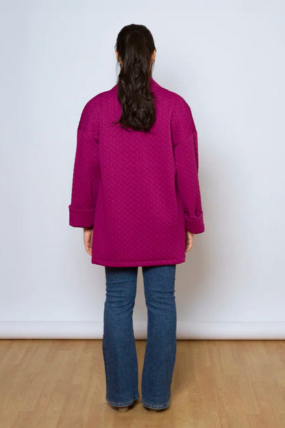 Back view of a woman wearing the Ina Coatigan by Tangente in Magenta, an open front cardigan in a cable-knit textured fabric, with dropped sleeves and patch pockets. She is wearing it with a white shirt and jeans and standing in front of a white background. 